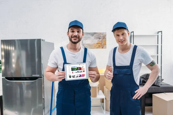 Two movers presenting digital tablet with aliexpress app on screen and showing thumb up sign in apartment — Stock Photo