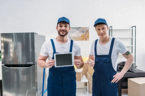 Due traslocatori che presentano tablet digitale con schermo vuoto, mostrando il segno pollice in alto e guardando la fotocamera in appartamento — Foto stock