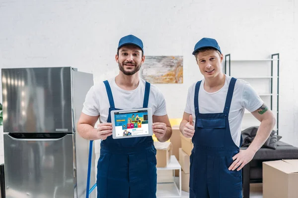 Two movers presenting digital tablet with amazon app on screen and showing thumb up sign in apartment — Stock Photo