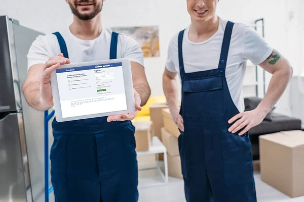 Cropped view of two movers presenting digital tablet with facebook app on screen in apartment — Stock Photo