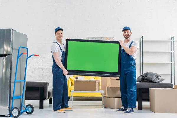 Dos personas en uniforme mirando a la cámara mientras transportan televisión con pantalla verde en el apartamento - foto de stock