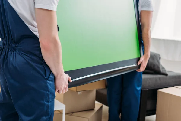 Cropped view of two movers in uniform transporting tv with green screen in apartment — Stock Photo