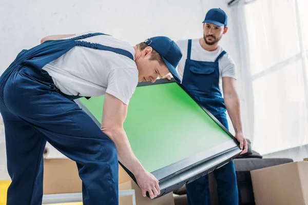 Zwei hübsche Mover in Uniform transportieren Fernseher mit grünem Bildschirm in der Wohnung — Stockfoto