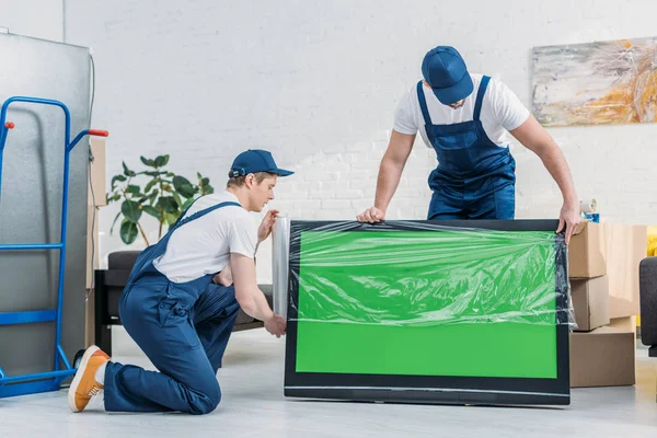Due traslocatori in uniforme utilizzando rotolo di film estensibile mentre avvolgono tv con schermo verde in appartamento — Foto stock