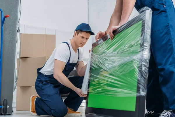 Two movers in uniform using roll of stretch film while wrapping tv with green screen in apartment — Stock Photo