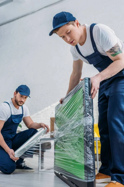 Two handsome movers in uniform using roll of stretch film while wrapping tv with green screen in apartment — Stock Photo