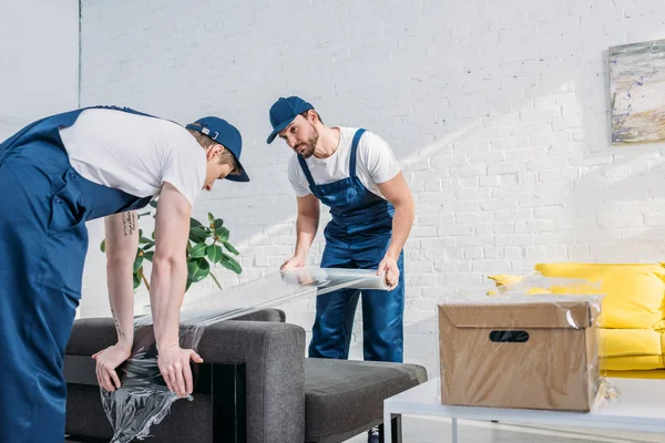 Déménageurs enveloppant canapé avec rouleau de film étirable dans l'appartement avec espace de copie — Photo de stock