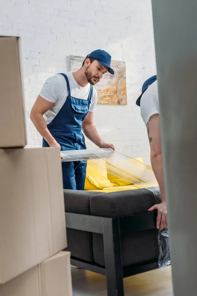 Selective focus of movers wrapping couch with roll of stretch film in apartment — Stock Photo