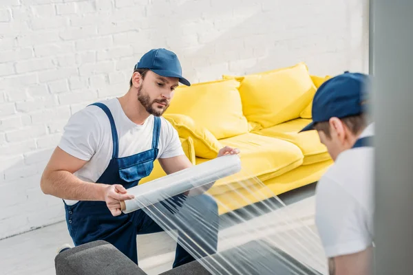 Movers in uniform wrapping furniture with roll of stretch film in apartment — Stock Photo