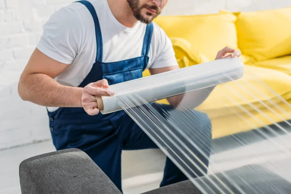 Cropped view of mover wrapping furniture with roll of stretch film in apartment — Stock Photo
