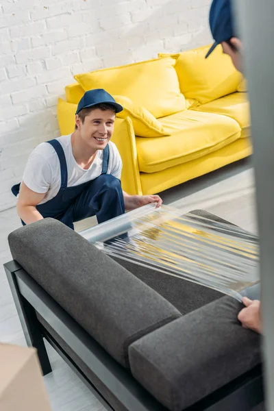 Canapé d'emballage souriant avec rouleau de film étirable dans l'appartement — Photo de stock