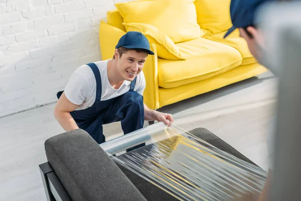 Smiling mover wrapping couch with roll of stretch film in apartment — Stock Photo