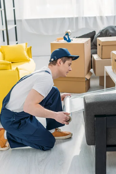 Déménageur en uniforme enveloppant des meubles avec rouleau de film étirable dans l'appartement — Photo de stock