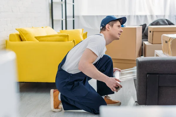 Foyer sélectif de déménageur dans des meubles d'emballage uniformes avec rouleau de film étirable dans l'appartement — Photo de stock