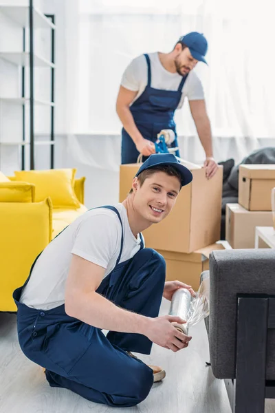 Movers wrapping furniture with roll of stretch film and packing boxes in apartment — Stock Photo