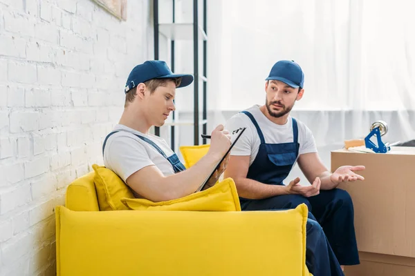 Two movers writing in clipboard and gesturing with hands while sitting on sofa in apartment — Stock Photo