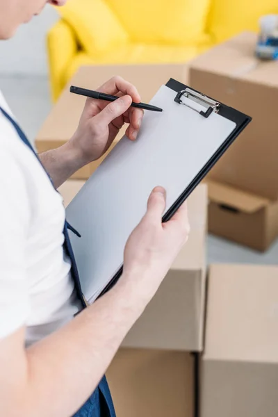 Cropped view of mover writing in empty clipboard near cardboard boxes in apartment — Stock Photo