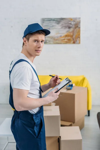 Beau déménageur tenant le presse-papiers et regardant la caméra près des boîtes en carton dans l'appartement — Photo de stock