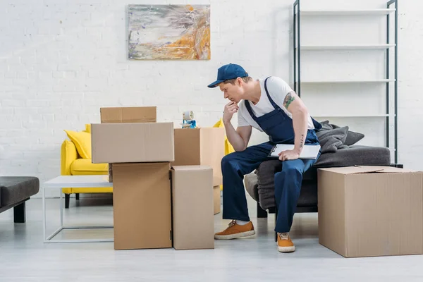 Pensive mover sitting near cardboard boxes and holding clipboard in apartment — Stock Photo