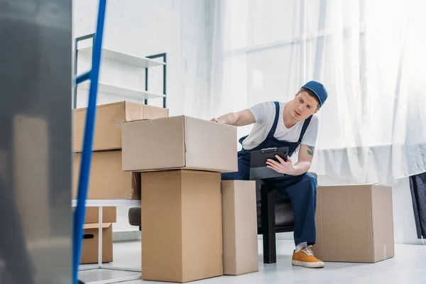 Déménageur assis près des boîtes en carton et tenant le presse-papiers dans l'appartement — Photo de stock