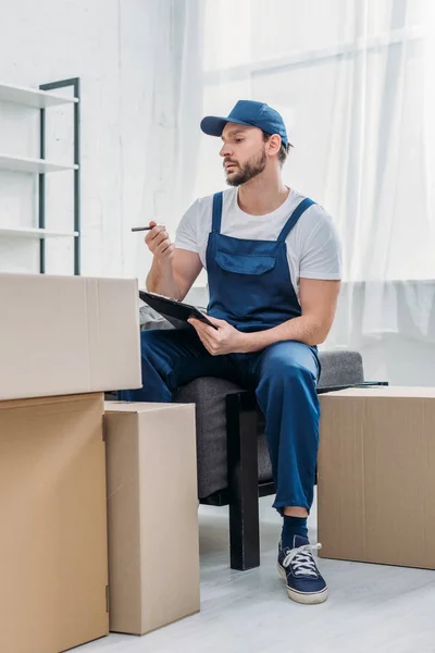 Schöner Mover sitzt neben Kartons und hält Klemmbrett in der Wohnung — Stockfoto