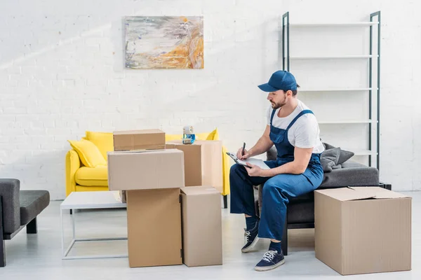 Handsome mover writing in clipboard while sitting near cardboard boxes in living room — Stock Photo