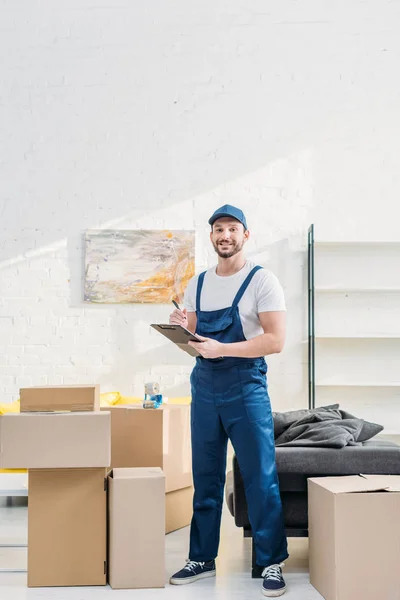 Mover looking at camera and holding clipboard near cardboard boxes in apartment with copy space — Stock Photo