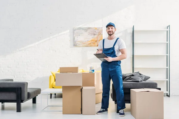 Mover blickt auf Kamera und hält Klemmbrett in der Nähe von Kartons in Wohnung mit Kopierraum — Stockfoto