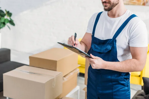 Cropped view of mover writing in clipboard near cardboard boxes in apartment — Stock Photo