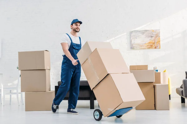 Mudanza en uniforme transportando cajas de cartón en camión de mano en apartamento - foto de stock