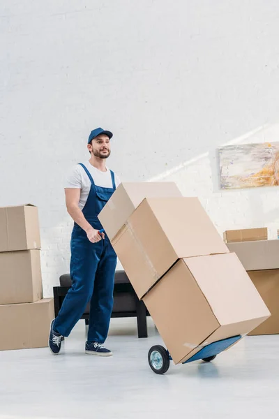 Mover in uniform transporting cardboard boxes on hand truck in apartment with copy space — Stock Photo