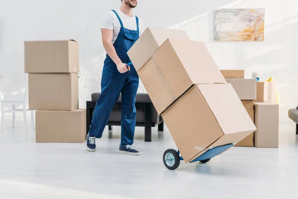 Vista cortada de mover em caixas de papelão de transporte uniforme no caminhão de mão no apartamento — Fotografia de Stock