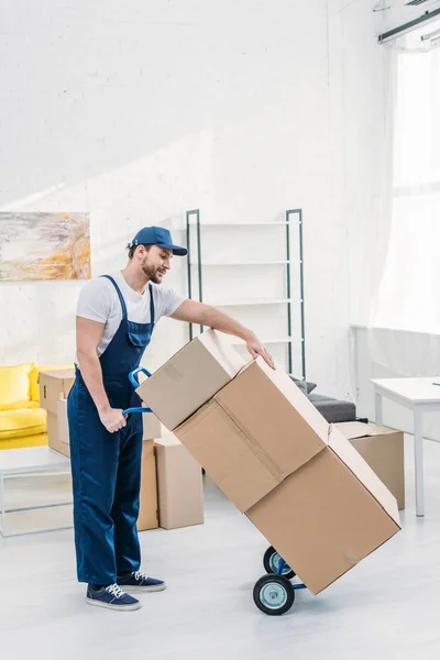 Déménageur en uniforme transportant des boîtes en carton sur camion à main dans l'appartement avec espace de copie — Photo de stock