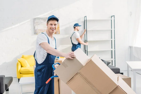 Two movers in uniform transporting cardboard boxes and furniture in apartment — Stock Photo