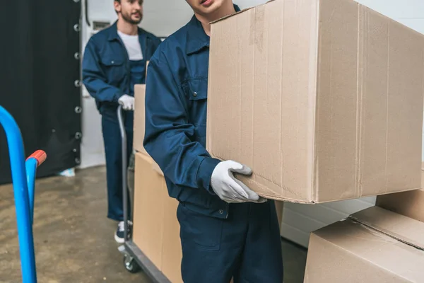 Vue recadrée de deux déménageurs en uniforme transportant des boîtes en carton avec camion à main dans l'entrepôt — Photo de stock