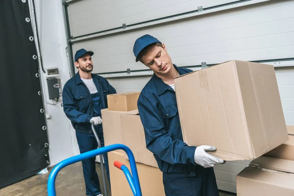 Two concentrated movers in uniform transporting cardboard boxes in warehouse — Stock Photo