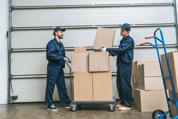 Two movers in uniform transporting cardboard boxes on hand truck in warehouse with copy space — Stock Photo