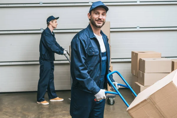 Deux déménageurs en uniforme transportant des boîtes en carton avec des camions à main dans l'entrepôt — Photo de stock