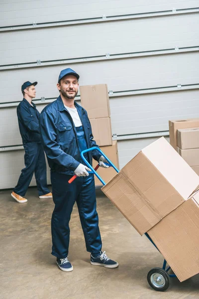 Deux déménageurs en uniforme transportant des boîtes en carton avec camion à main dans l'entrepôt — Photo de stock