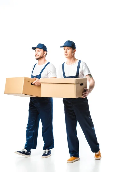 Deux beaux déménageurs en uniforme transportant des boîtes en carton sur blanc — Photo de stock