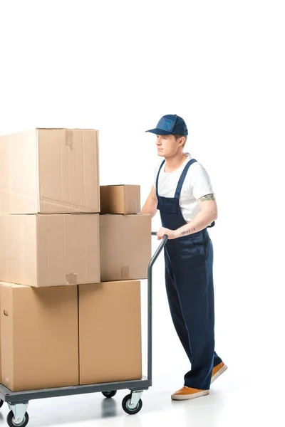 Handsome mover in uniform transporting cardboard boxes on hand truck isolated on white — Stock Photo