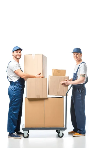 Deux beaux déménageurs regardant la caméra tout en transportant des boîtes en carton avec camion à main sur blanc — Photo de stock