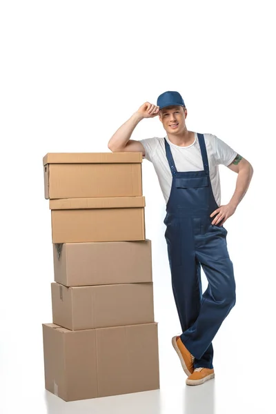 Beau déménageur en uniforme avec des boîtes en carton regardant la caméra tout en ajustant le chapeau isolé sur blanc — Photo de stock