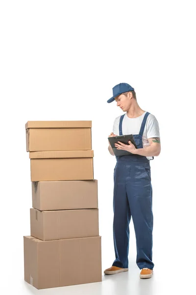Vista parcial del guapo transportista en escritura uniforme en portapapeles cerca de cajas de cartón aisladas en blanco - foto de stock