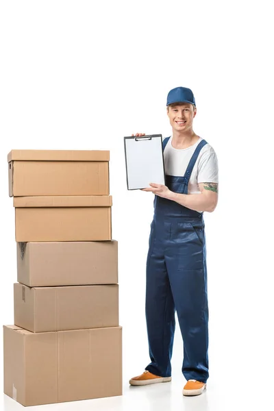 Smiling mover in uniform looking at camera and holding blank clipboard near cardboard boxes isolated on white — Stock Photo
