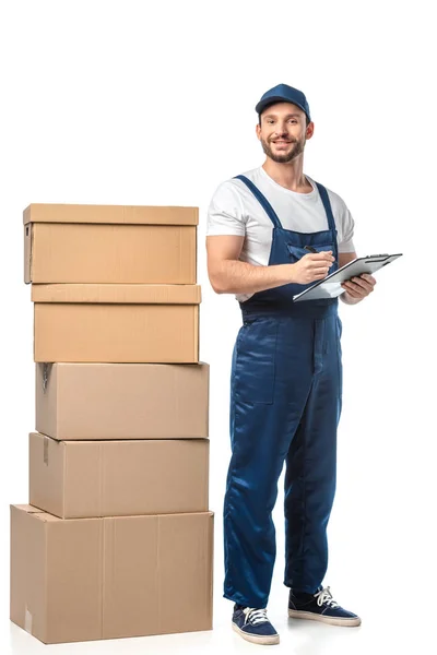 Déménageur en uniforme regardant la caméra tout en écrivant dans le presse-papiers près de boîtes en carton isolé sur blanc — Photo de stock