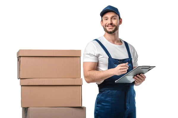 Mover en uniforme mirando a la cámara mientras se escribe en portapapeles cerca de cajas de cartón aisladas en blanco - foto de stock