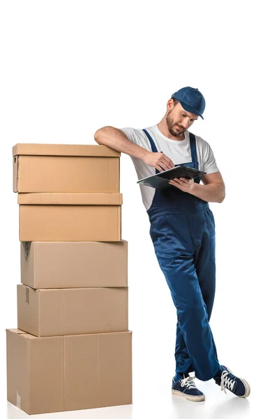 Mover in uniform writing in clipboard near cardboard boxes isolated on white — Stock Photo