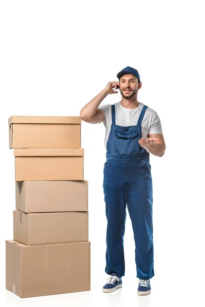 Handsome smiling mover with cardboard boxes talking on smartphone and gesturing with hand isolated on white — Stock Photo