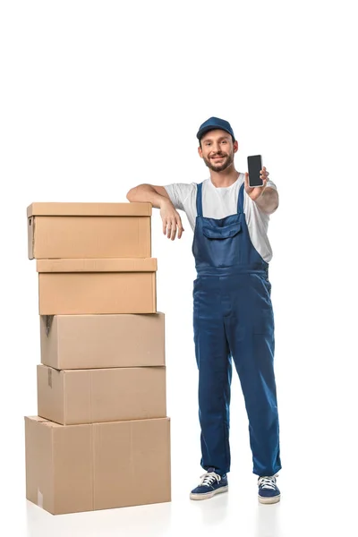 Handsome smiling mover with cardboard boxes presenting smartphone with blank screen isolated on white — Stock Photo